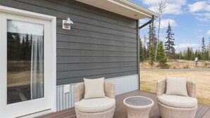 A deck with two wicker chairs and a small table, adjacent to a building with dark siding and a white window.