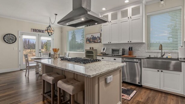 A modern kitchen with white cabinets, a central island with bar stools, stainless steel appliances, and a large window with blinds.