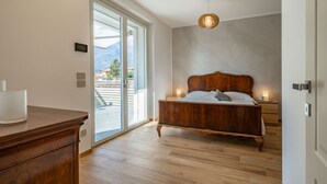 A bedroom with a wooden bed, a nightstand, a bedside lamp, and a view of mountains through a sliding door. - Villa Sunshine - Luxury property with large garden and Pool, near the lake, parking e-charge, ideal for people with mobility issues (Dervio)