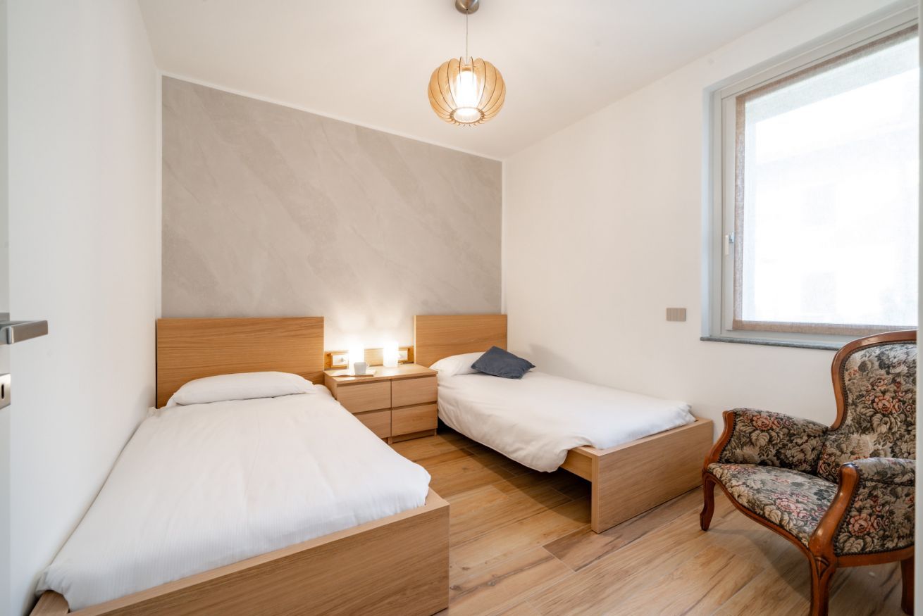 Une chambre avec deux lits simples, une table de chevet en bois avec une lampe, une chaise et une grande fenêtre.