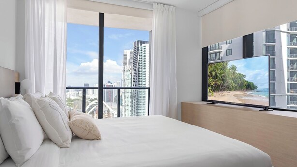 A modern hotel room with a large bed, white linens, a flat-screen TV, and a balcony with a city view. - Chic 2-bedroom apartment in vibrant Miami (Miami)