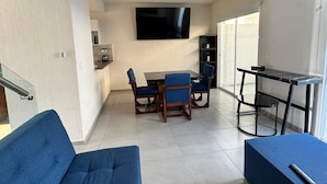 A modern living room with a blue sofa, a dining table with chairs, a flat-screen TV mounted on the wall, and a small kitchen area with cabinets and a countertop. - Residencia Altavela Privada 51 (Valle de Banderas)
