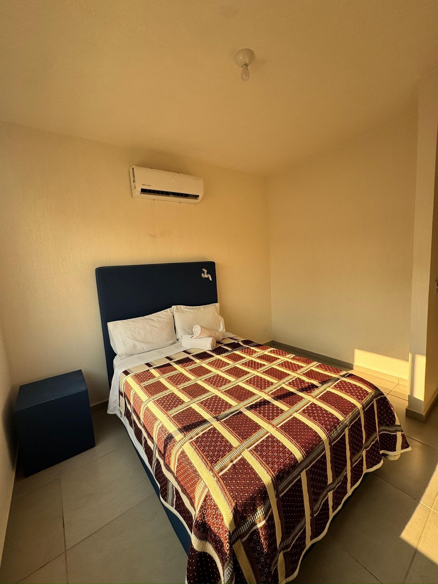 A hotel room with a bed, a pillow, a blanket, a bedside table, and an air conditioning unit on the wall.