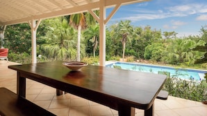 Uma área externa coberta com piscina, uma mesa com um recipiente e vegetação exuberante ao fundo.