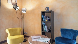 Um quarto com duas cadeiras, uma mesinha, uma estante e um abajur.