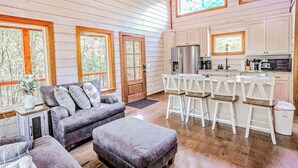 L’intérieur chaleureux d’un chalet comprend un espace de vie avec un canapé et un pouf, une cuisine dotée de meubles blancs et d’un réfrigérateur, ainsi que de grandes fenêtres offrant une vue sur la forêt environnante.