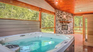 Un bain à remous dans un chalet en bois, doté d’une cheminée en pierre et de grandes fenêtres donnant sur un paysage verdoyant.