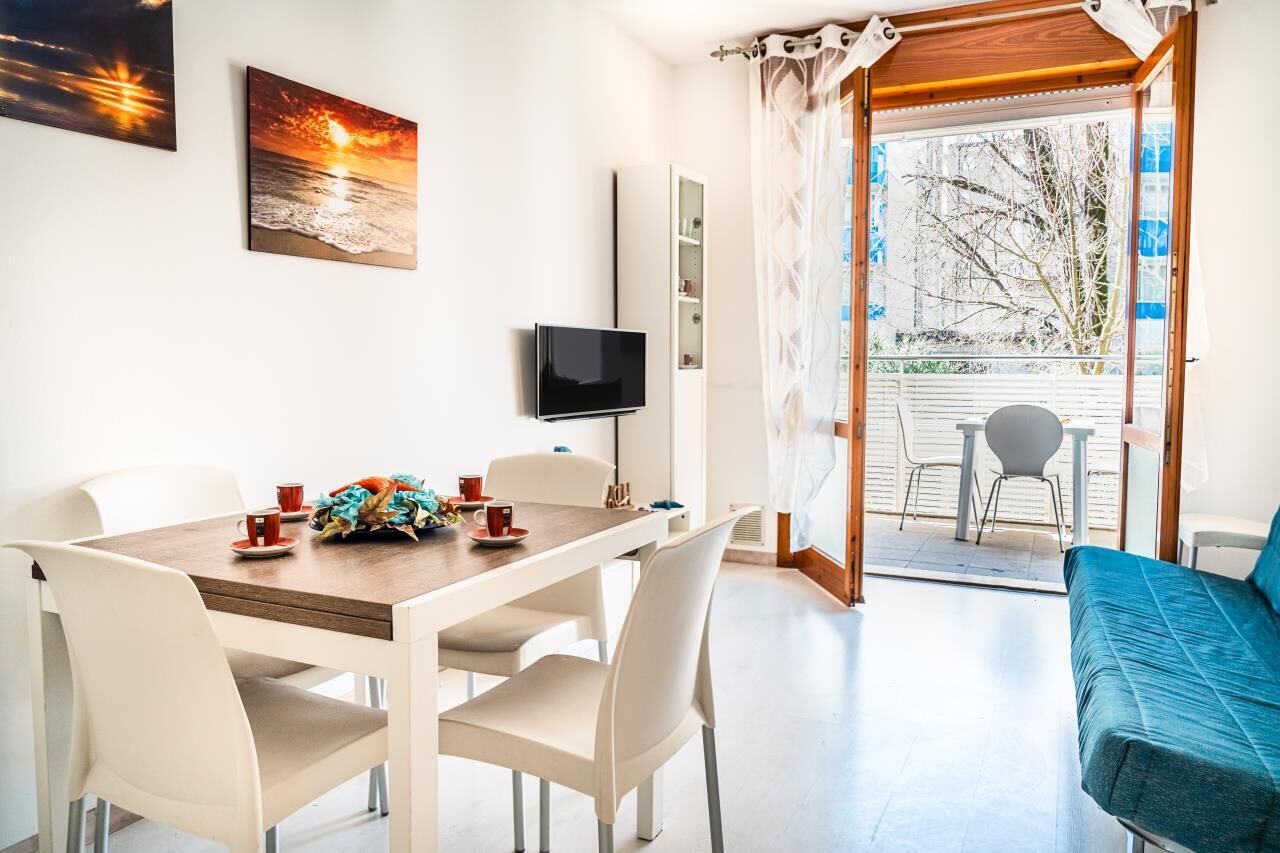 A dining area with a table set for two, a television mounted on the wall, and a balcony with a view of trees and a fence.