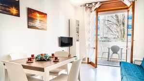 Een eetgedeelte met een tafel voor twee personen, een aan de muur bevestigde televisie, en een balkon met uitzicht op bomen en een hek.
