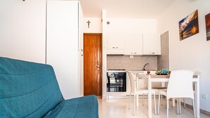Een compacte leefruimte met een blauwgroene bank, een eethoek met witte stoelen, een keuken met witte kastjes en een houten deur.