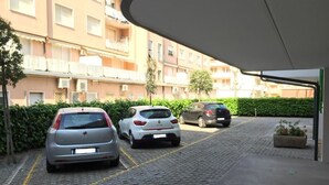 Ein Parkplatz mit drei nebeneinander stehenden Autos, ein Gebäude mit Balkonen im Hintergrund und ein Hecke im Vordergrund.