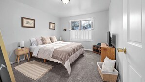 Quarto com cama, mesas de cabeceira, televisão, janela com persianas e porta com puxador.