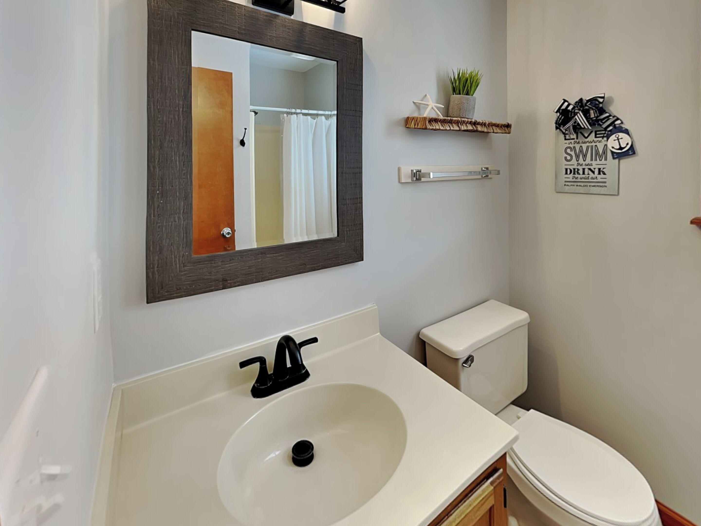 A bathroom with a white sink, a toilet, a mirror, and a shelf with a plant and a sign that says "SWIM for the sea, drink for the sea."