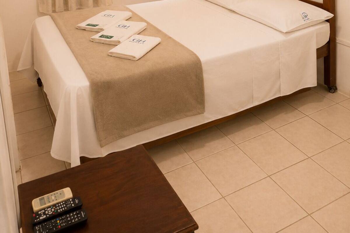 Una habitación de hotel con cama, mesita de noche con lámpara, ventana con cortinas, aire acondicionado instalado en la pared y un cuadro enmarcado.