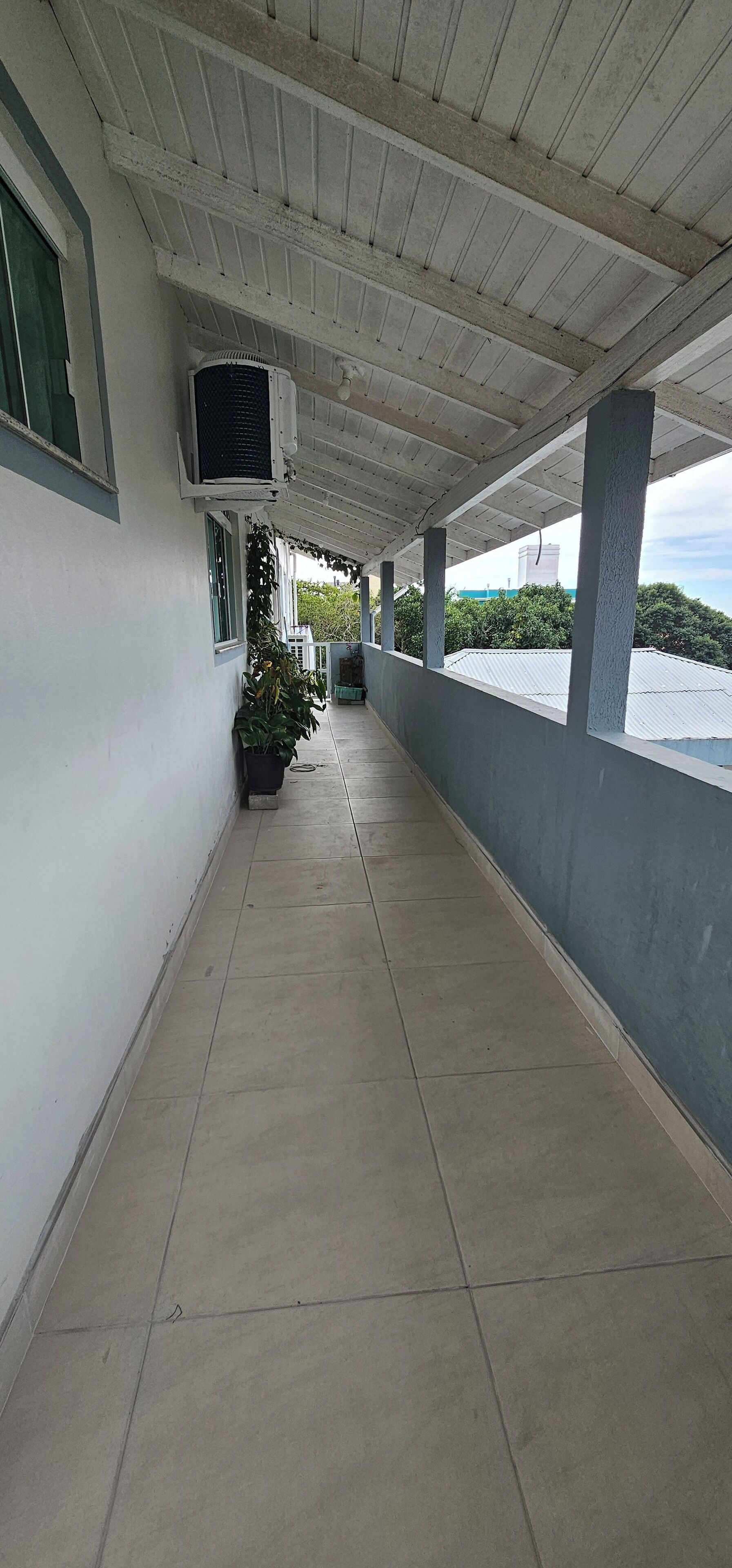 A covered corridor with tiled flooring, air conditioning units, potted plants, and a view of greenery and a distant building.