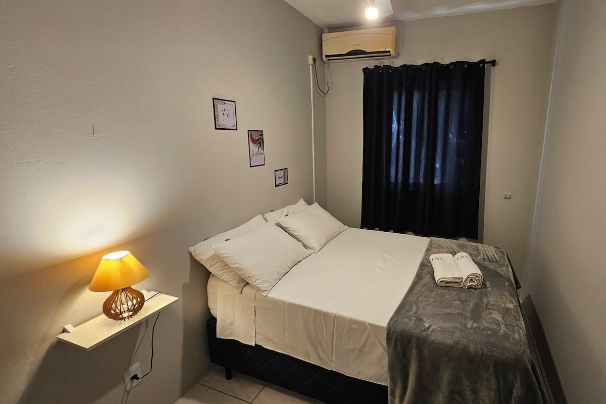 Un dormitorio con cama, lámpara de noche, una mesita, ventana con cortinas y un aire acondicionado.