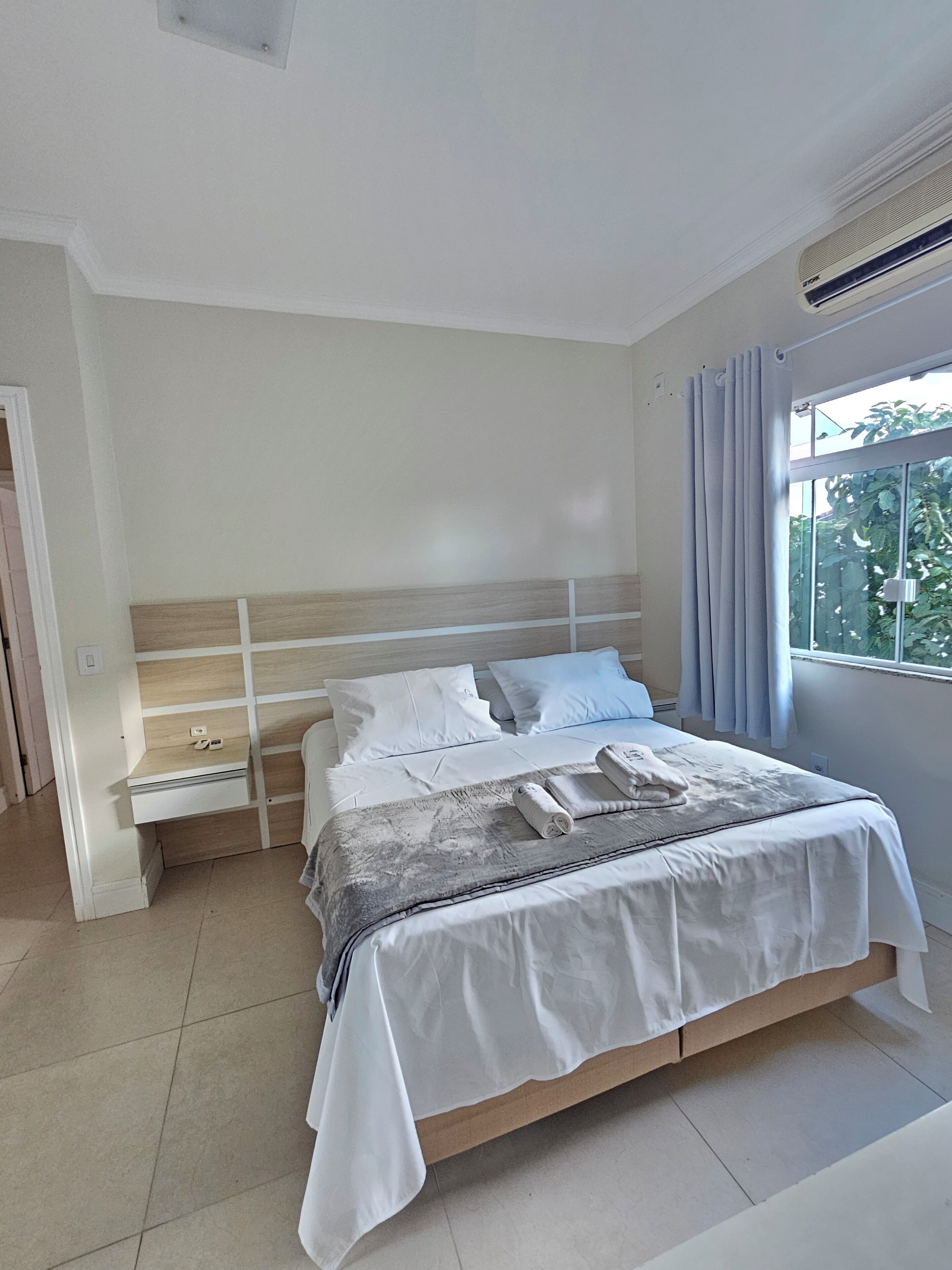 Une chambre moderne avec une tête de lit en bois, du linge de lit blanc, une table de chevet avec un téléphone et une fenêtre avec des rideaux.