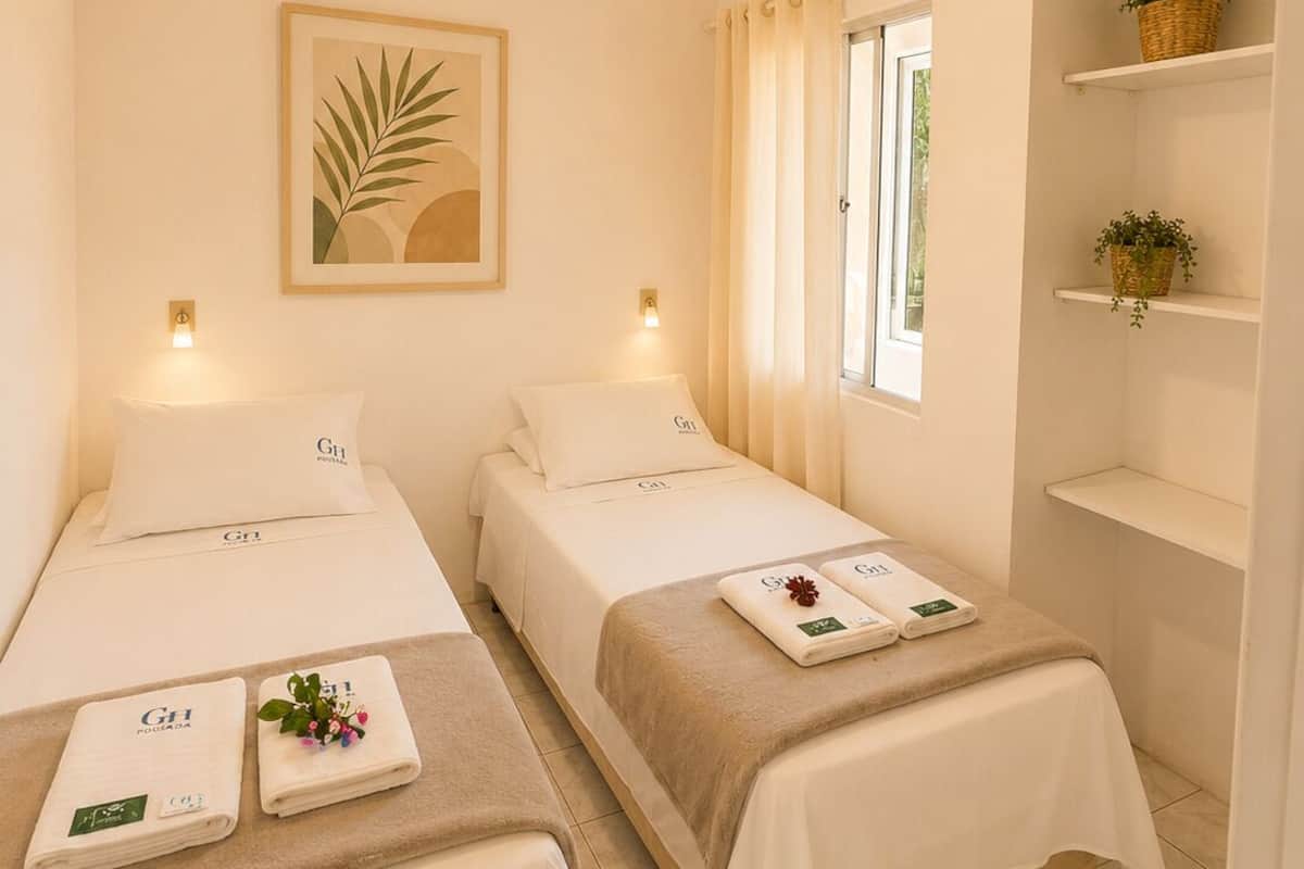 Habitación de hotel con dos camas, ventilador de techo, ventana con cortinas y una repisa en la pared con plantas.