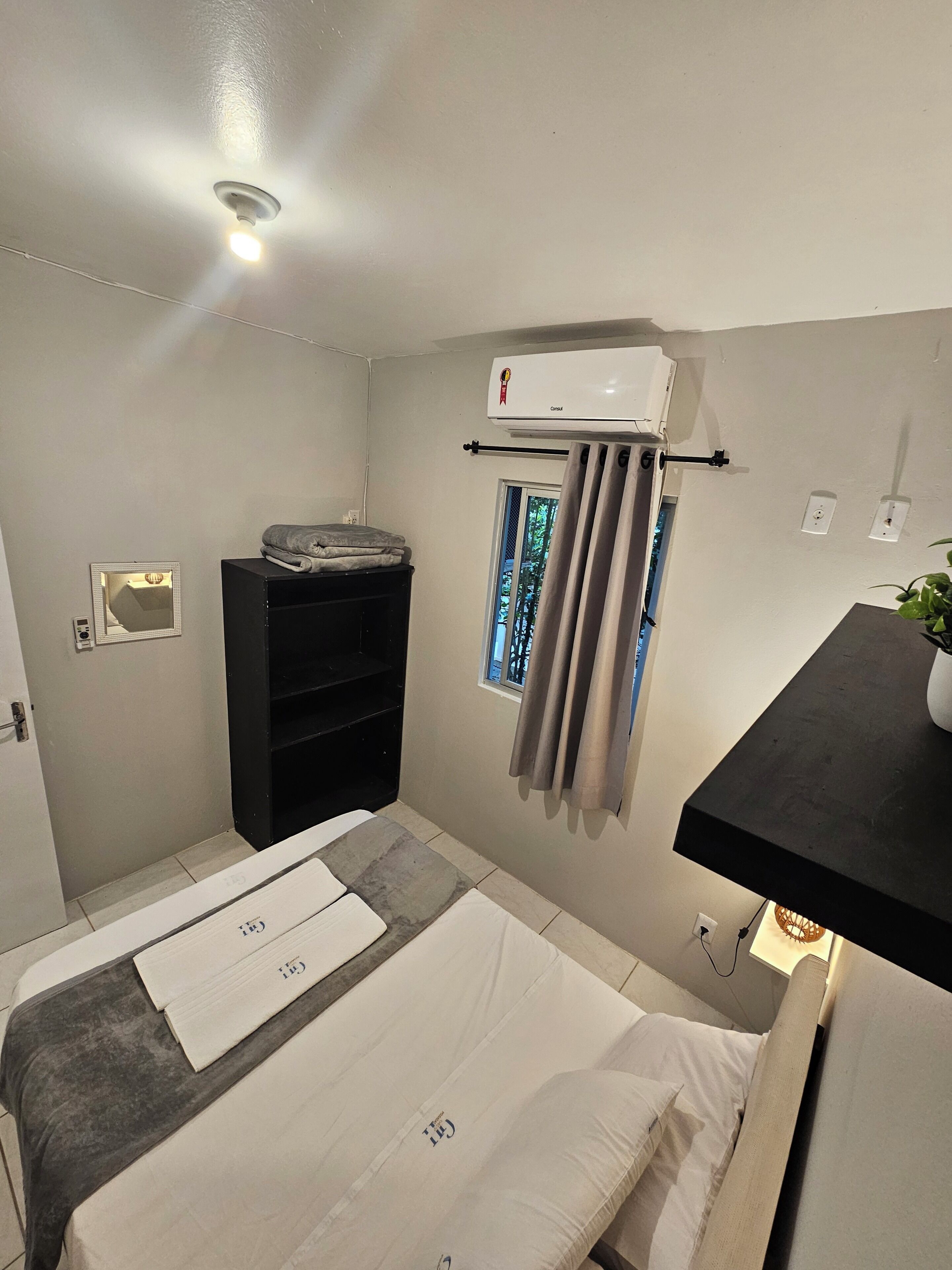 A compact hotel room with a bed, a small desk, a window with curtains, an air conditioning unit, and a wall-mounted shelf.