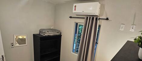 A compact hotel room with a bed, a small desk, a window with curtains, an air conditioning unit, and a wall-mounted shelf.