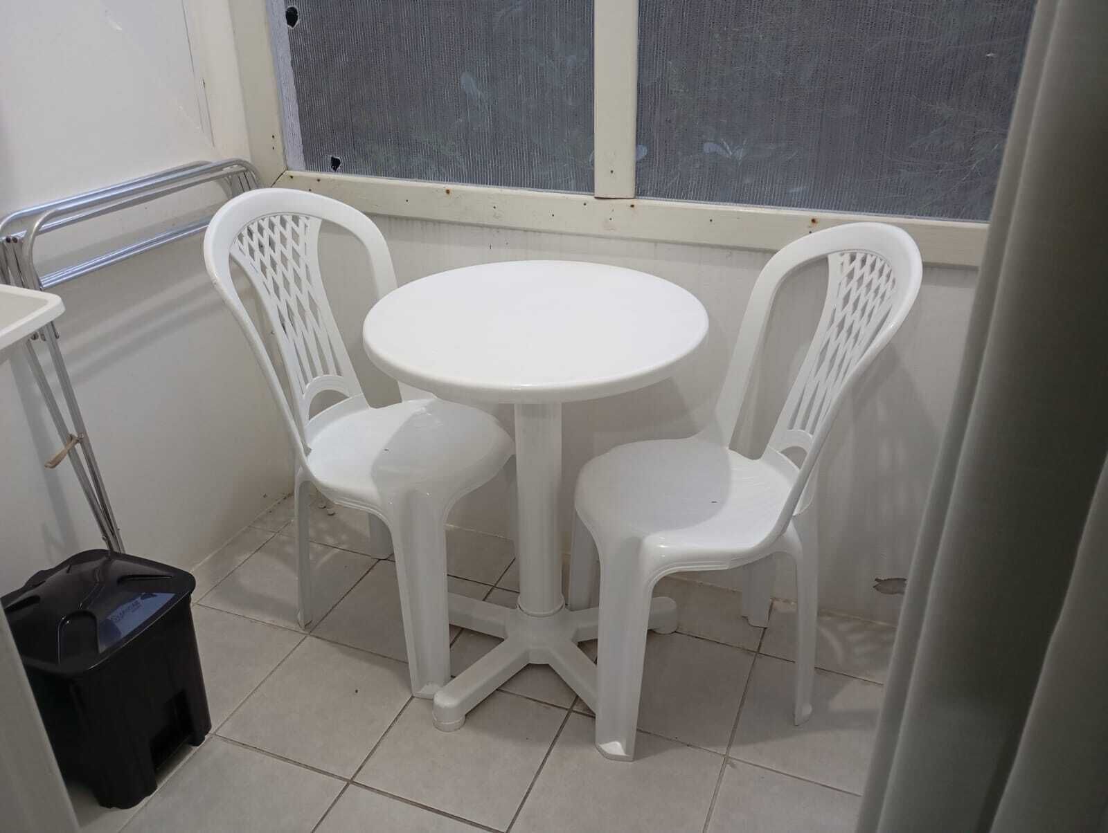 Una habitación pequeña con una mesa redonda y dos sillas, un cubo de basura negro y una ventana con mosquitero.