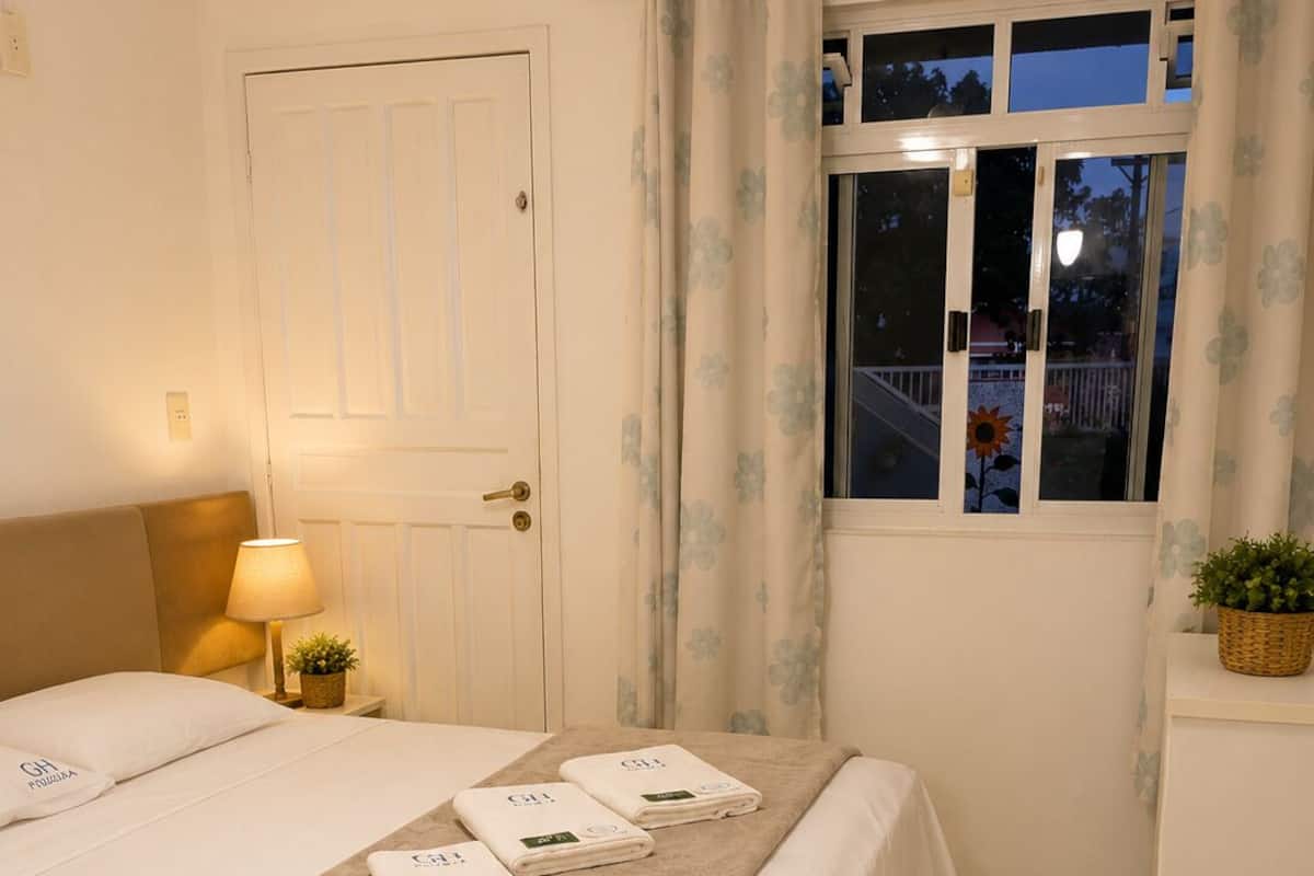 Habitación de hotel con cama, mesita de noche con lámpara, ventana con cortinas y vistas al exterior.