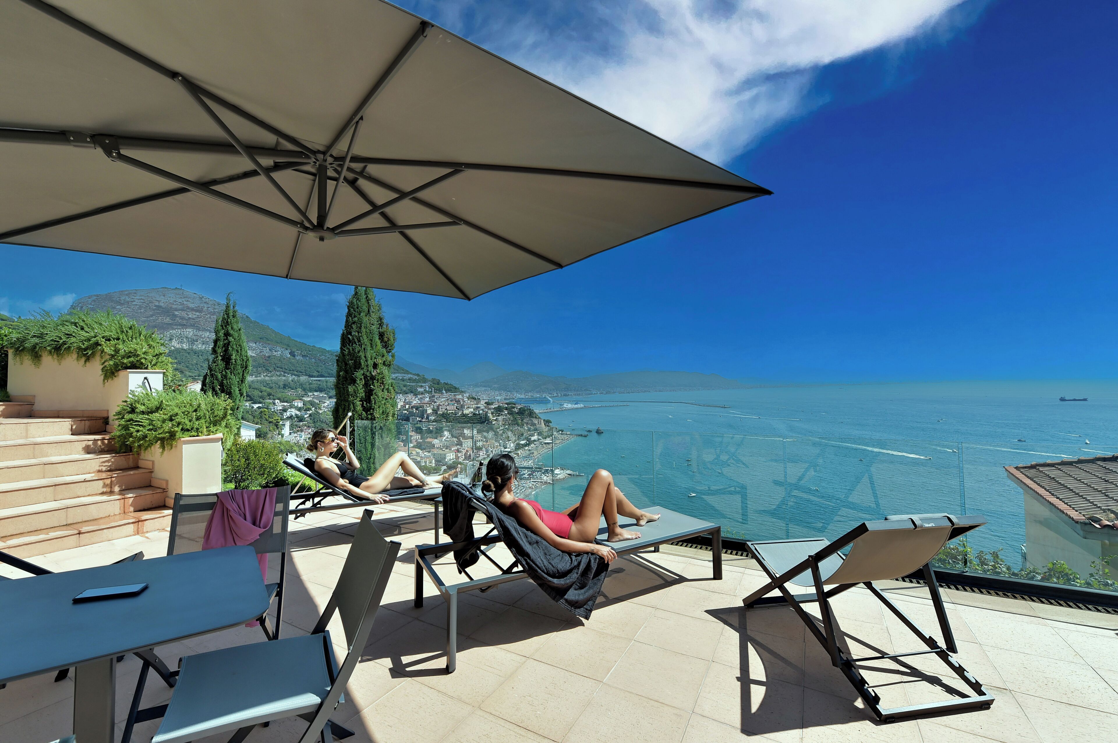A terrace with lounge chairs and a large umbrella, overlooking a coastal city and the sea.