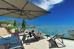A terrace with lounge chairs and a large umbrella, overlooking a coastal city and the sea.