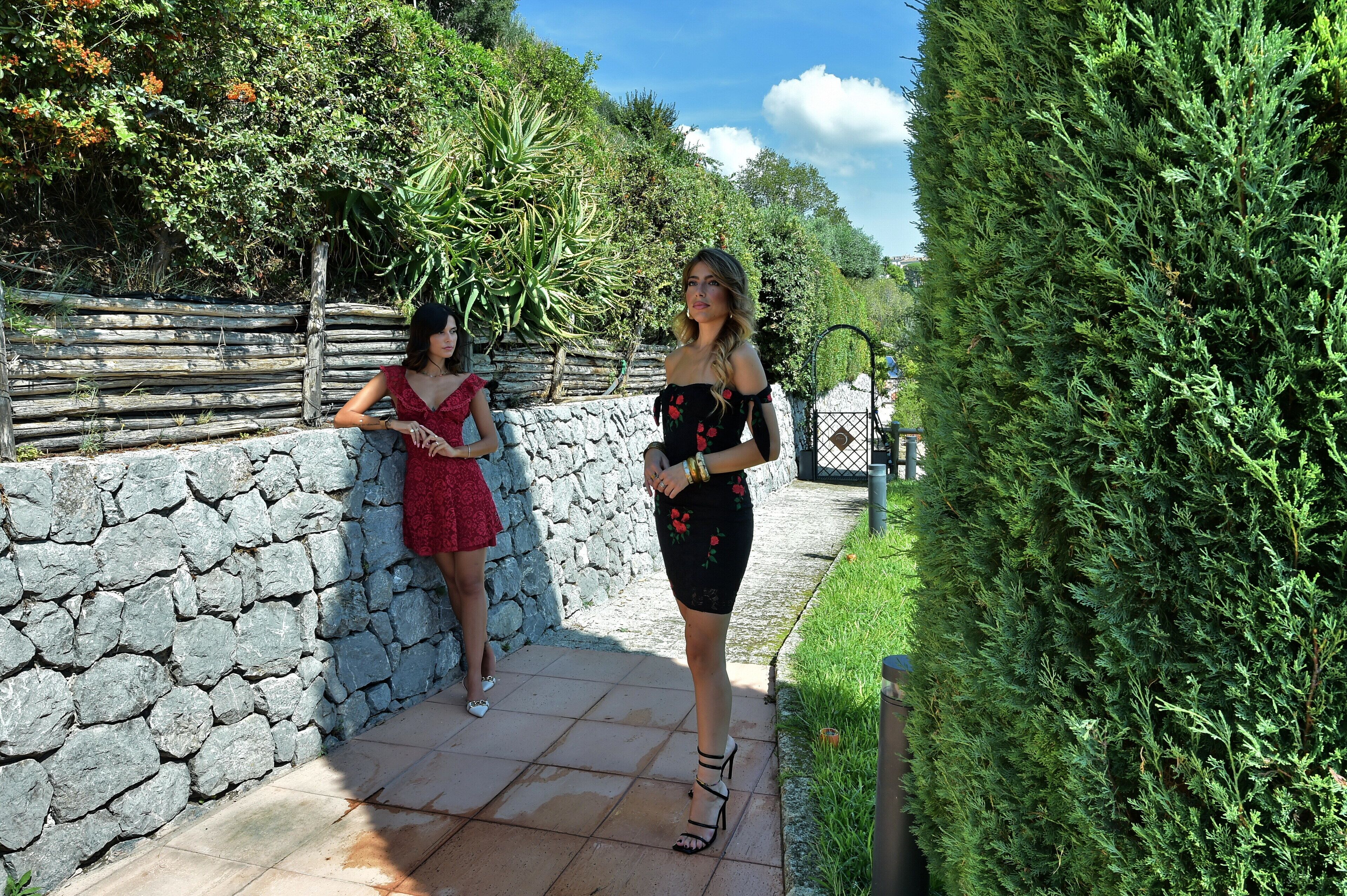Two women standing on a stone pathway with a stone wall and greenery on one side and a hedge on the other.