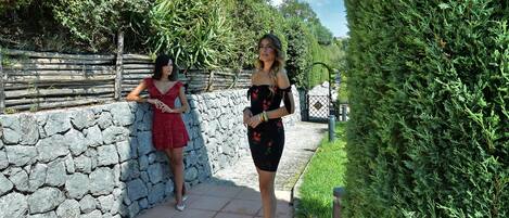 Two women standing on a stone pathway with a stone wall and greenery on one side and a hedge on the other.