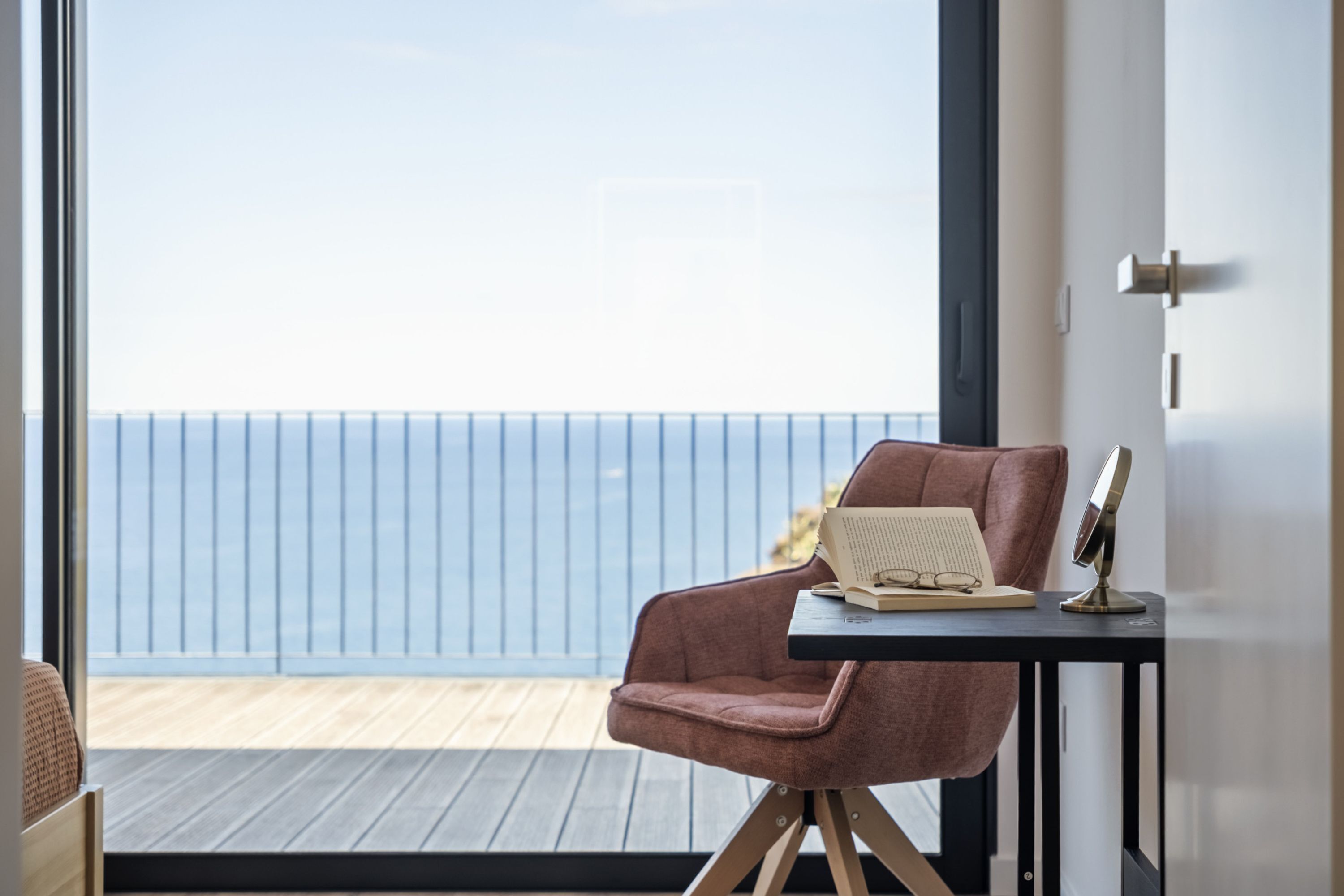 A room with a view of the sea, a pink chair, a small table with a book and glasses, and a mirror.