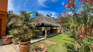 Un jardín tropical con una estructura de techo de paja, plantas en macetas y un cielo azul claro.