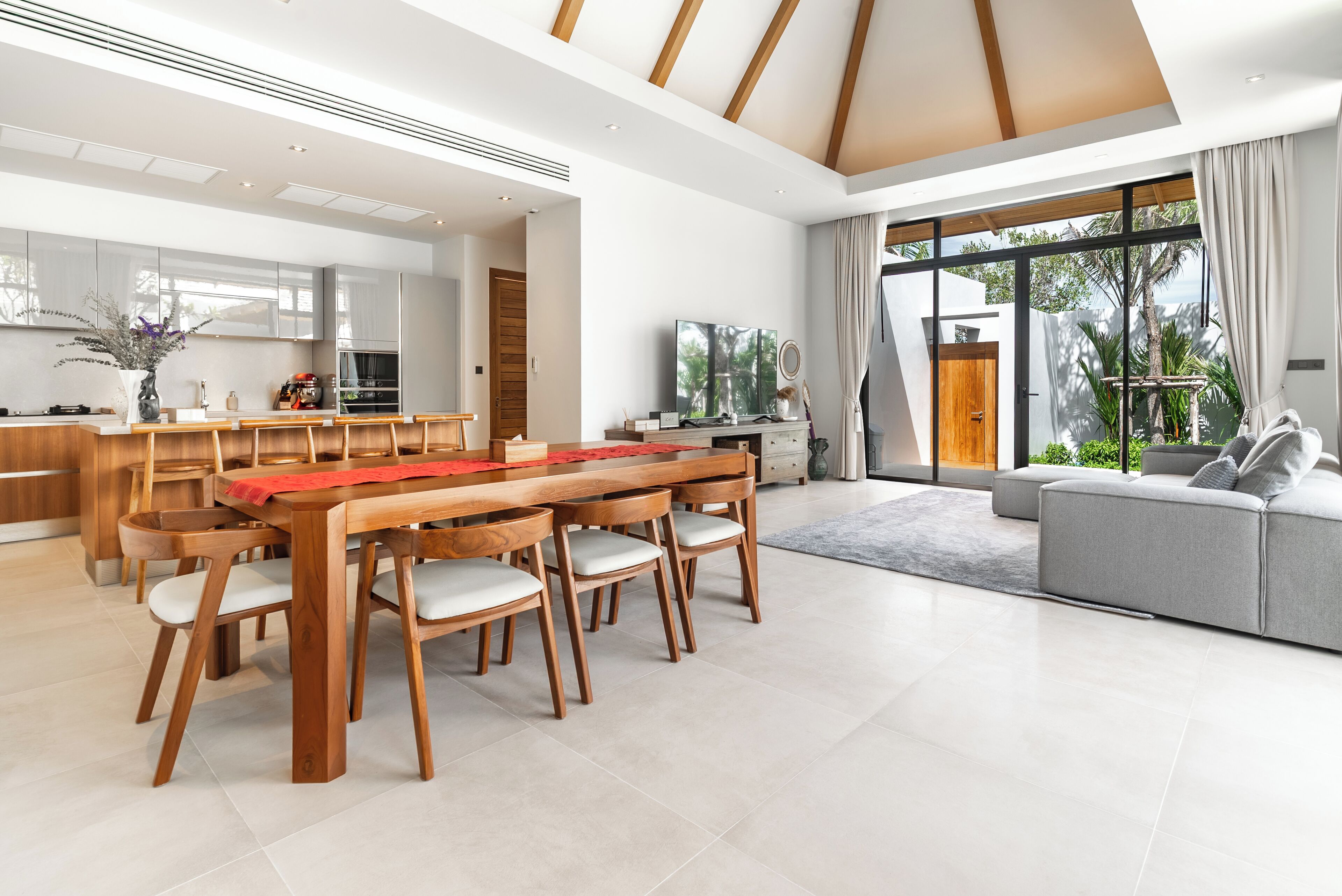 Una cocina moderna con armarios de madera, un comedor con mesa y sillas de madera, un sofá gris y ventanas amplias con vistas a zonas verdes.