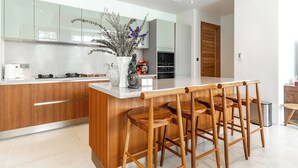 A modern kitchen with wooden cabinets, a central island with bar stools, a stainless steel refrigerator, and a potted plant. - Anchan Flora By Villacarte (Thep Krasattri)