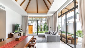 A spacious living area with a wooden dining table, a sofa, and a large window offering a view of the outdoors. - Anchan Flora By Villacarte (Thep Krasattri)