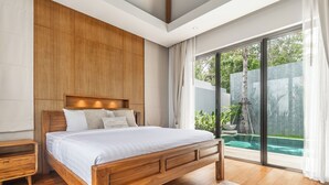 A modern bedroom with a wooden bed, a nightstand, and a large sliding glass door leading to a pool area. - Anchan Flora By Villacarte (Thep Krasattri)