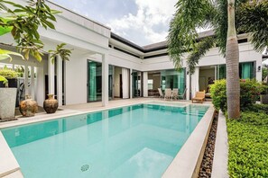 Uma área de piscina moderna com piscina transparente, rodeada por um edifício branco com portas de vidro e um alpendre coberto.