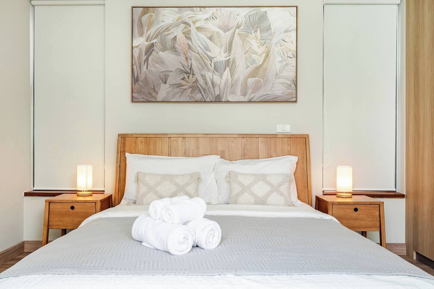 Ein ordentlich bezogenes Bett mit weißen Leintüchern und zusammengerollten Handtüchern, begleitet von zwei Nachttischen mit Lampen, in einem Raum mit einem Holz-Kopfteil und einem großen floralen Kunstwerk darüber.