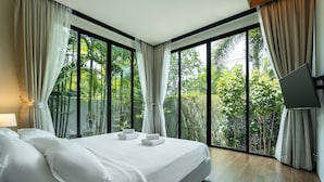Une chambre moderne avec un grand lit, du linge de lit blanc et une vue sur une végétation luxuriante grâce à de grandes fenêtres du sol au plafond.