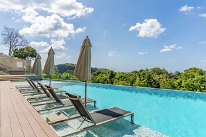Zona junto a la piscina con sillones de descanso y sombrillas, rodeada de exuberante vegetación y un cielo despejado.