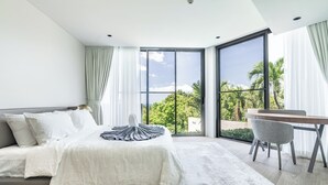 Ein modernes Schlafzimmer mit einem großen Bett, einem Schreibtisch und Blick auf Palmen durch raumhohe Fenster.