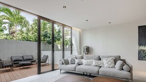 Ein modernes Wohnzimmer mit einem grauen Sofa, einem Holztisch und einer Glastür, die auf einen Balkon mit Pflanzen führt.