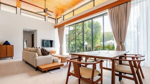 Una sala de estar moderna con un sofá, una mesa de comedor de madera con sillas, un ventilador de techo y ventanas grandes con cortinas.