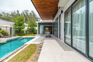 A modern outdoor pool area with a wooden ceiling, a glass-enclosed lounge, and a stone pathway. - Botanica Modern By Villacarte 159/32 (Si Sunthon)
