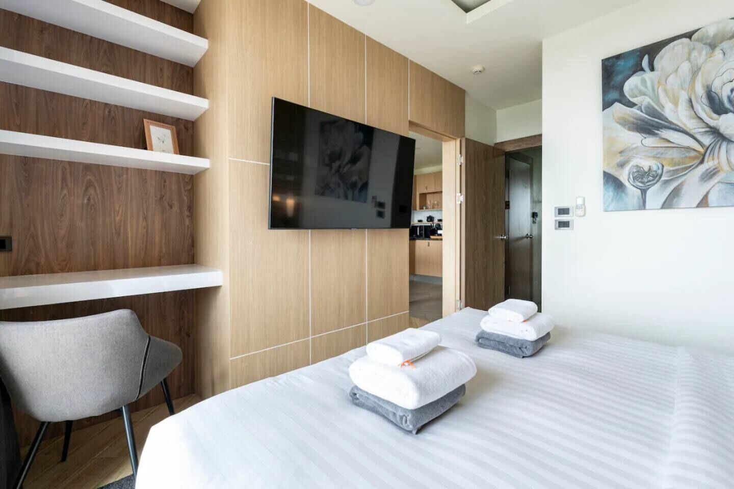 A modern hotel room with a large bed, a flat-screen TV, a painting, and a view into the kitchen area.