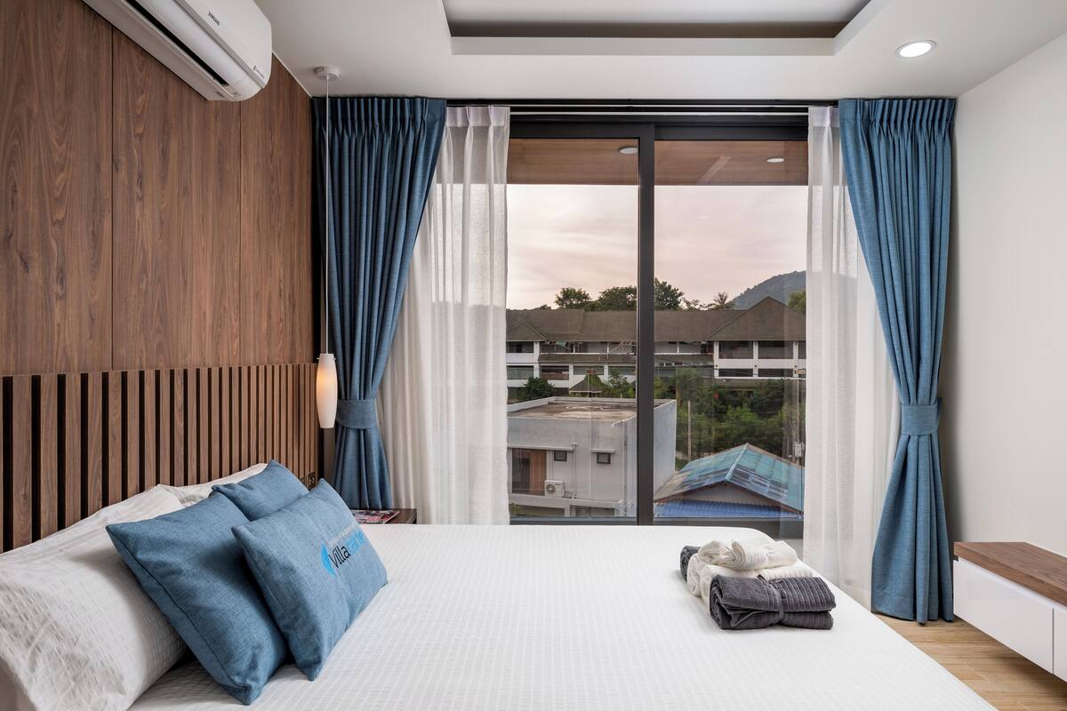 A modern hotel room with a large bed, a wooden headboard, a view through a glass door, and a ceiling light.