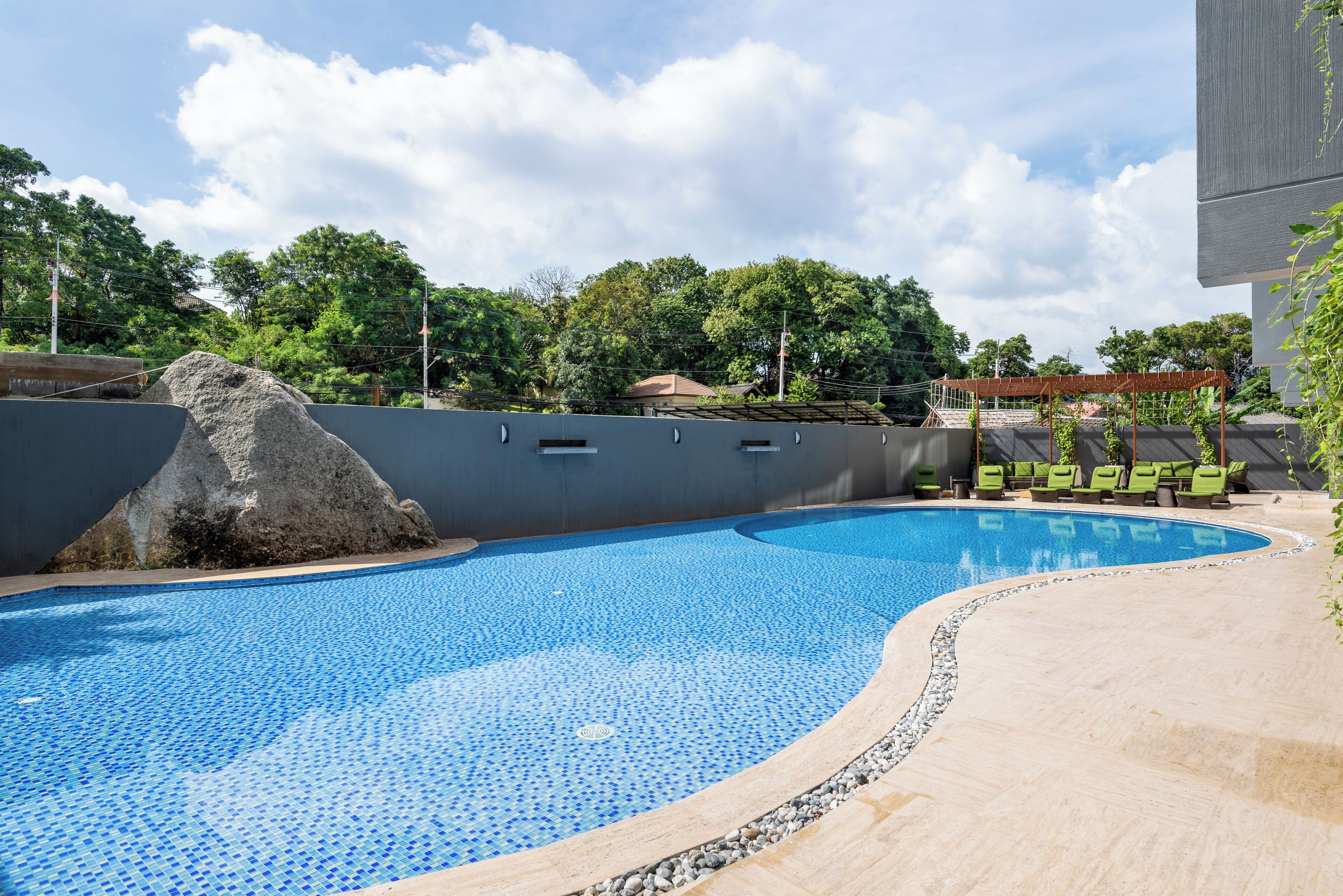 Una piscina con piso de baldosas, un elemento decorativo de roca y una pérgola con sillas.