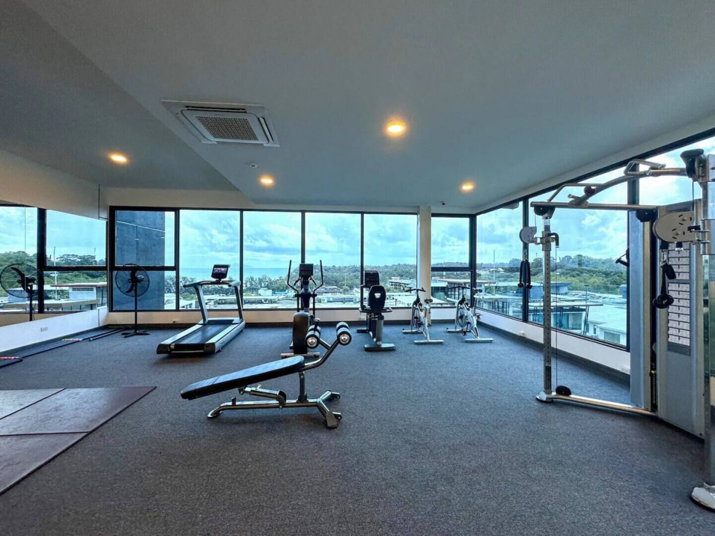 Un gimnasio con máquinas de cardio, pesas libres y una prensa de pesas. Amplias ventanas ofrecen vistas del paisaje urbano.