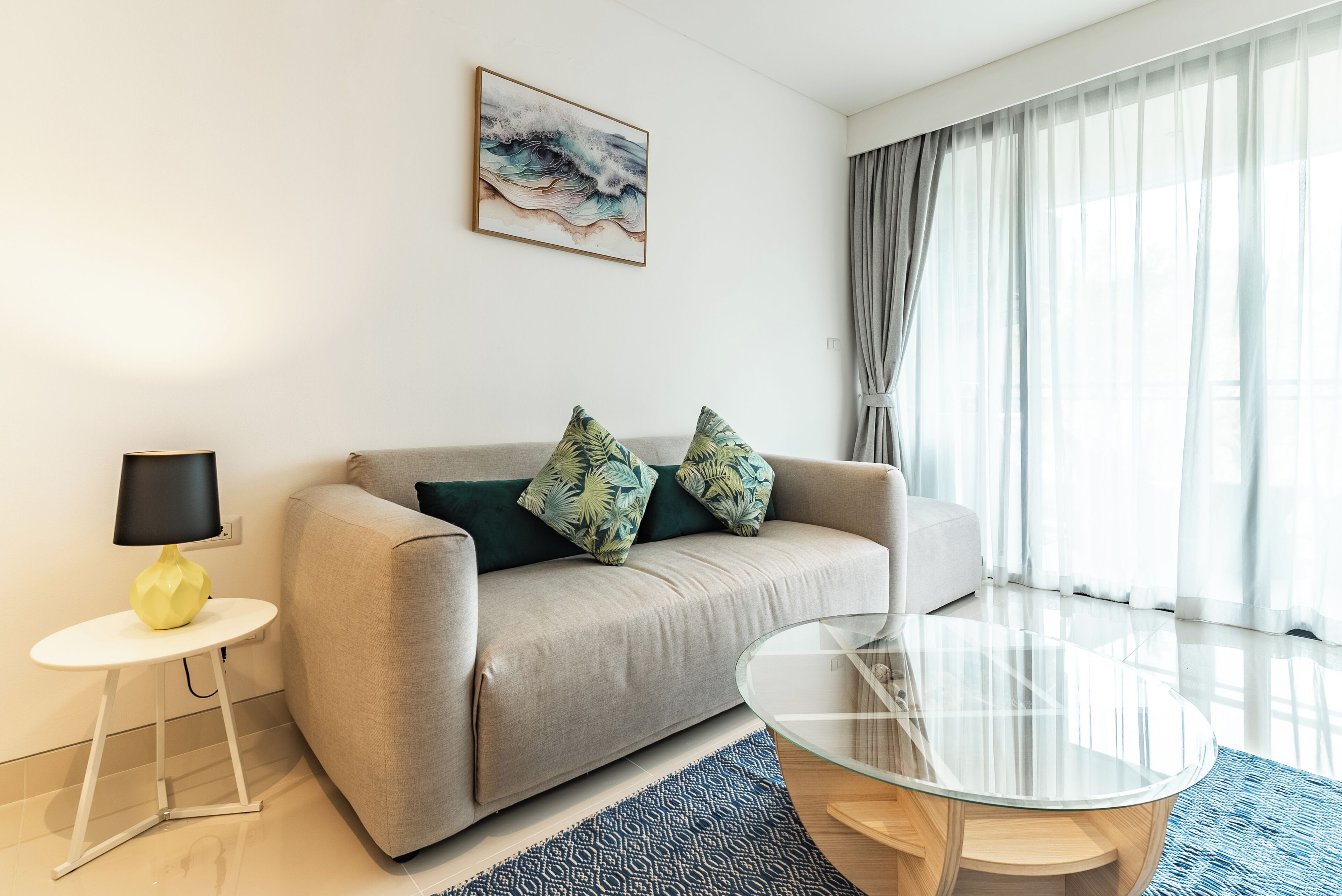A modern living room with a sofa, a glass coffee table, a lamp, and a painting on the wall.
