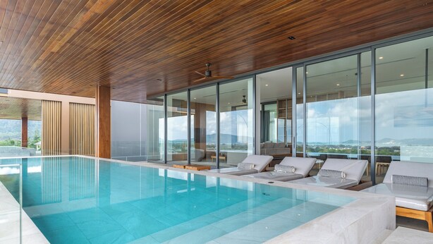 A modern indoor swimming pool with clear blue water, wooden ceiling, and large glass windows offering a view of the outdoors. - La vista Villas by Villacarte (Chalong)